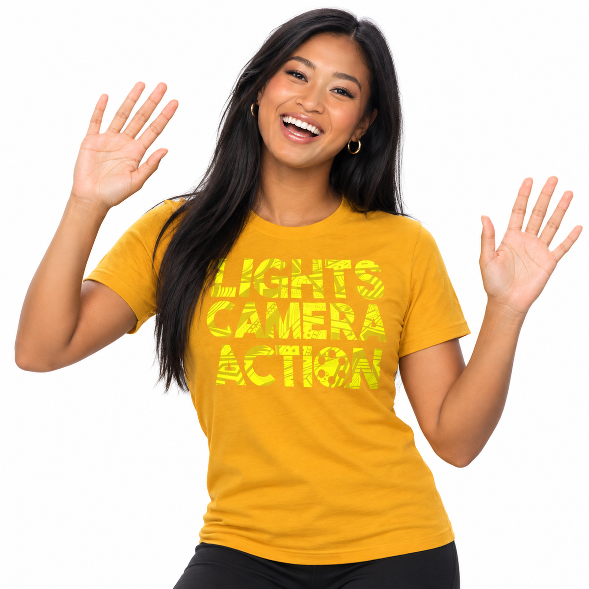 A woman with long black hair smiling and waving, wearing a mustard yellow graphic t-shirt that reads 'LIGHTS CAMERA ACTION' in a distressed yellow font, set against a plain white background.