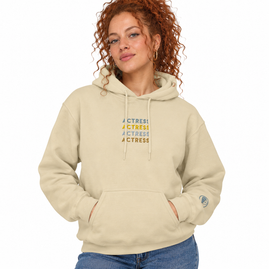 A woman with curly red hair wearing a beige pullover hoodie with 'ACTRESS' printed in blue and yellow on the chest, paired with blue jeans, against a plain white background.