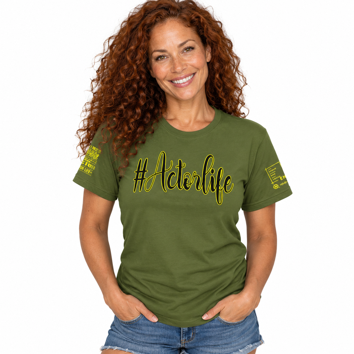 A smiling woman with long curly red hair wearing an olive green graphic t-shirt with yellow text reading '#Actorlife' across the chest, paired with denim shorts, against a plain white background.