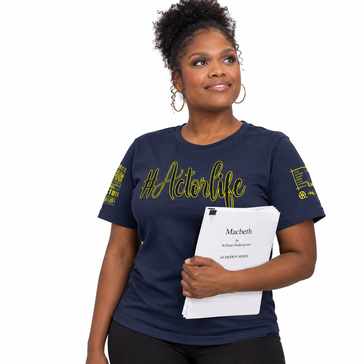 A woman modeling a navy blue graphic t-shirt with yellow text reading '#ActorLife' and sleeve prints, holding a script book against a plain white background.