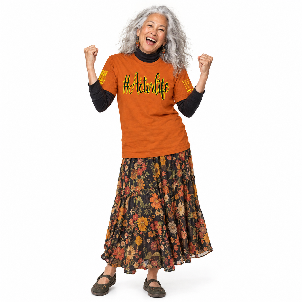 An older woman with long gray hair is smiling and celebrating with her fists raised, wearing an orange graphic t-shirt with black sleeves, a black turtleneck, a long black maxi skirt with a colorful floral print, and dark brown shoes, against a plain white background.