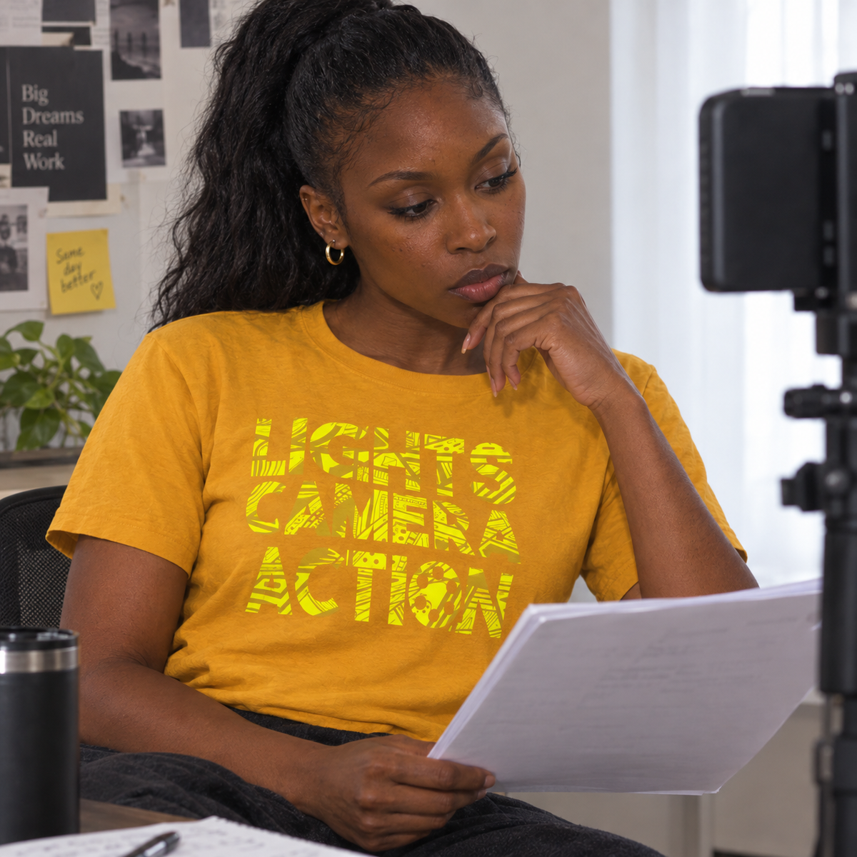 A woman wearing a bright yellow graphic t-shirt with the words 'LIGHTS CAMERA ACTION' in distressed yellow lettering, sitting in an office setting and looking at a document.