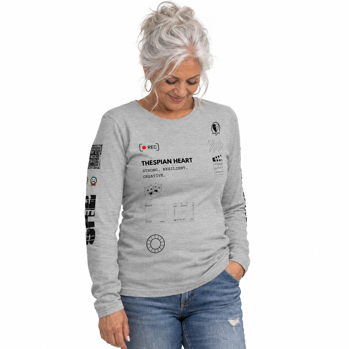 A woman with silver hair looking down, wearing a light gray long-sleeve t-shirt with black graphic text and logos, paired with blue jeans, against a plain white background.
