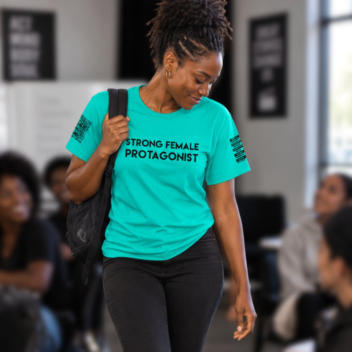 A woman walking in an indoor event space wearing a bright turquoise graphic T-shirt with the words 'STRONG FEMALE PROTAGONIST' printed in black across the chest, paired with black pants and carrying a black shoulder bag.