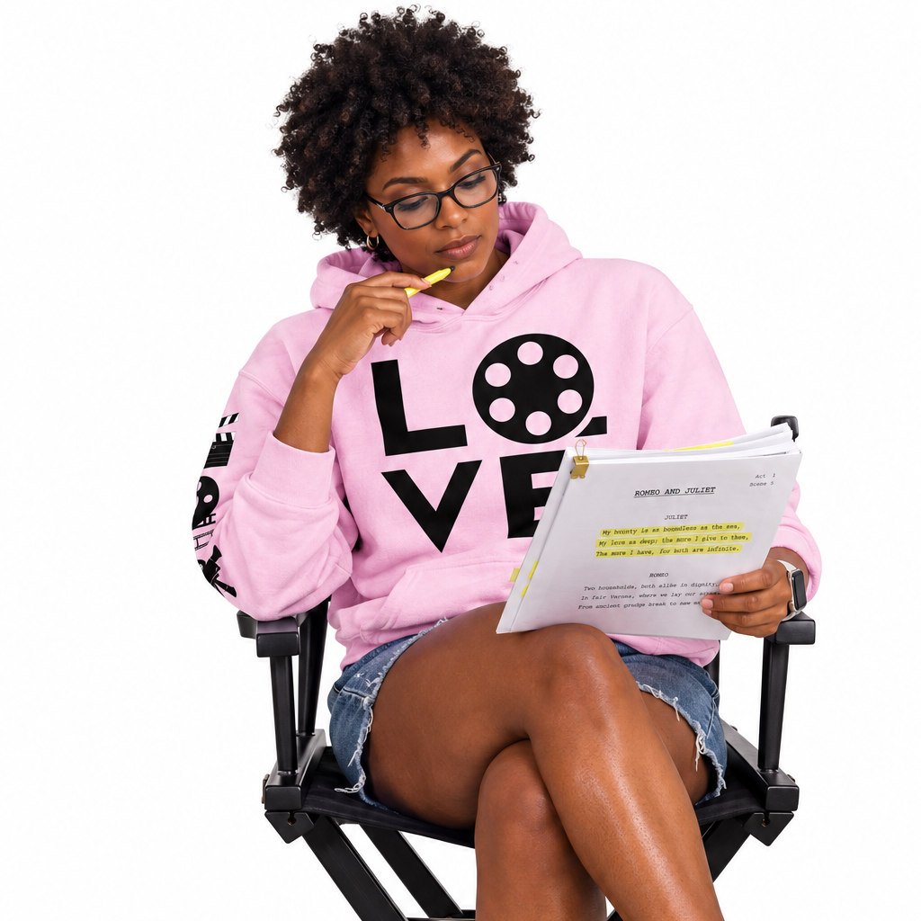 A woman with curly hair and glasses sits in a black director chair, reading a script. She is wearing a pink hoodie with 'LOVE' printed on the front, denim shorts, and white sneakers.
