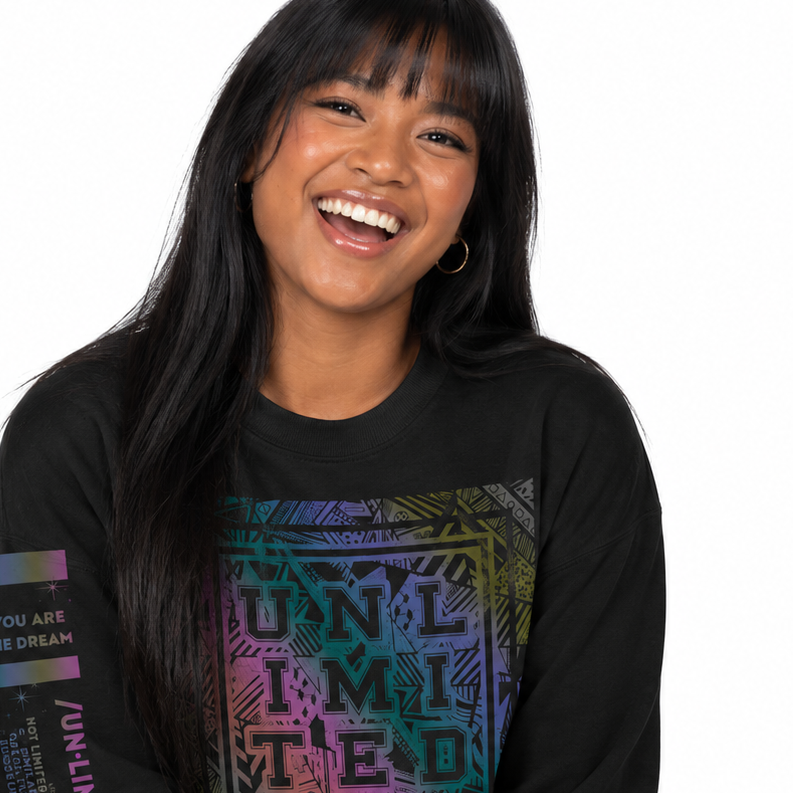 A woman smiling while wearing a black long-sleeve graphic sweatshirt with a colorful 'UNLIMITED' print on the front and a multicolor sleeve graphic, paired with blue jeans against a white background.