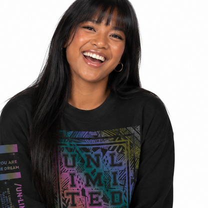 A woman smiling while wearing a black long-sleeve graphic sweatshirt with a colorful 'UNLIMITED' print on the front and a multicolor sleeve graphic, paired with blue jeans against a white background.