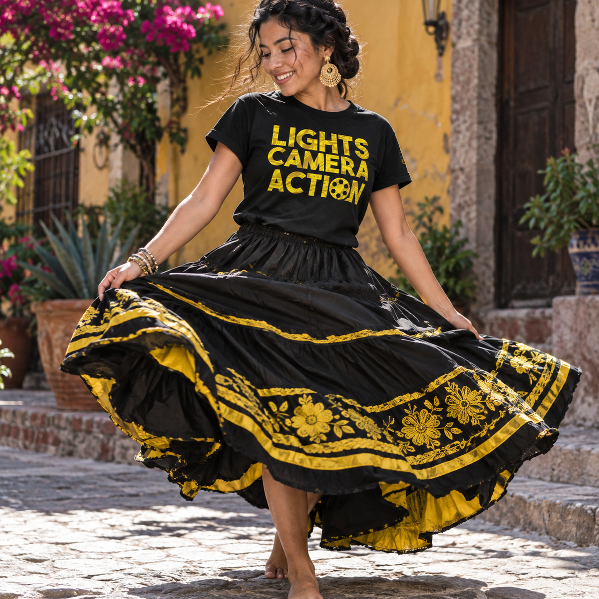 A woman outdoors wearing a black graphic T-shirt that reads 'LIGHTS CAMERA ACTION' in yellow and a voluminous black skirt with bold yellow floral and border embroidery. She is twirling with the skirt spread wide in a cobblestone street setting with a yellow wall and flowers in the background.